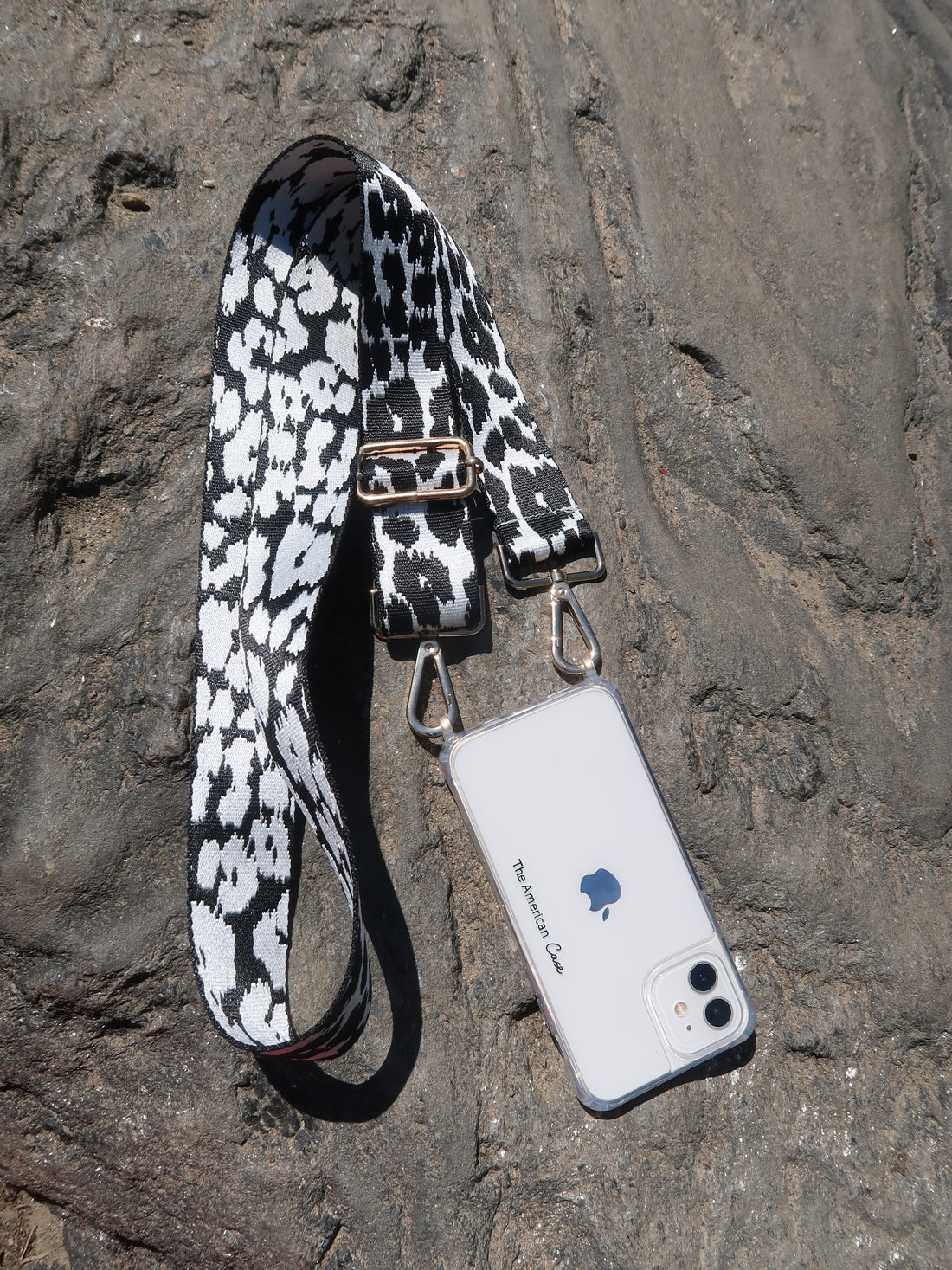 Adjustable black and white leopard Print Strap with Golden Carabiners attached to a clear The American Case phone case on a rock