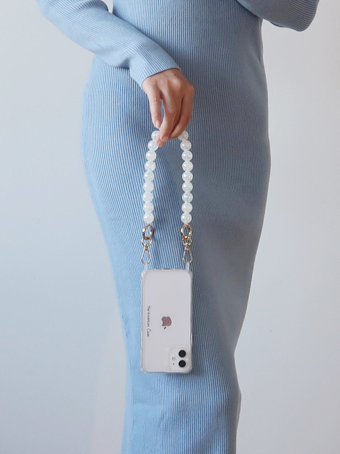 a Pastel white Crackle Bead Short Phone Chain with Golden Carabiners attache to a clear phone case with The American Case print hold by a lady by her hand
