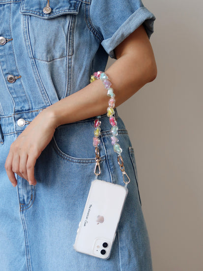 A short bead chain with golden carabiners and a glittery heart charm in pink, blue, yellow, and purple colors. The chain is attached to a phone hold by a lady on her hand