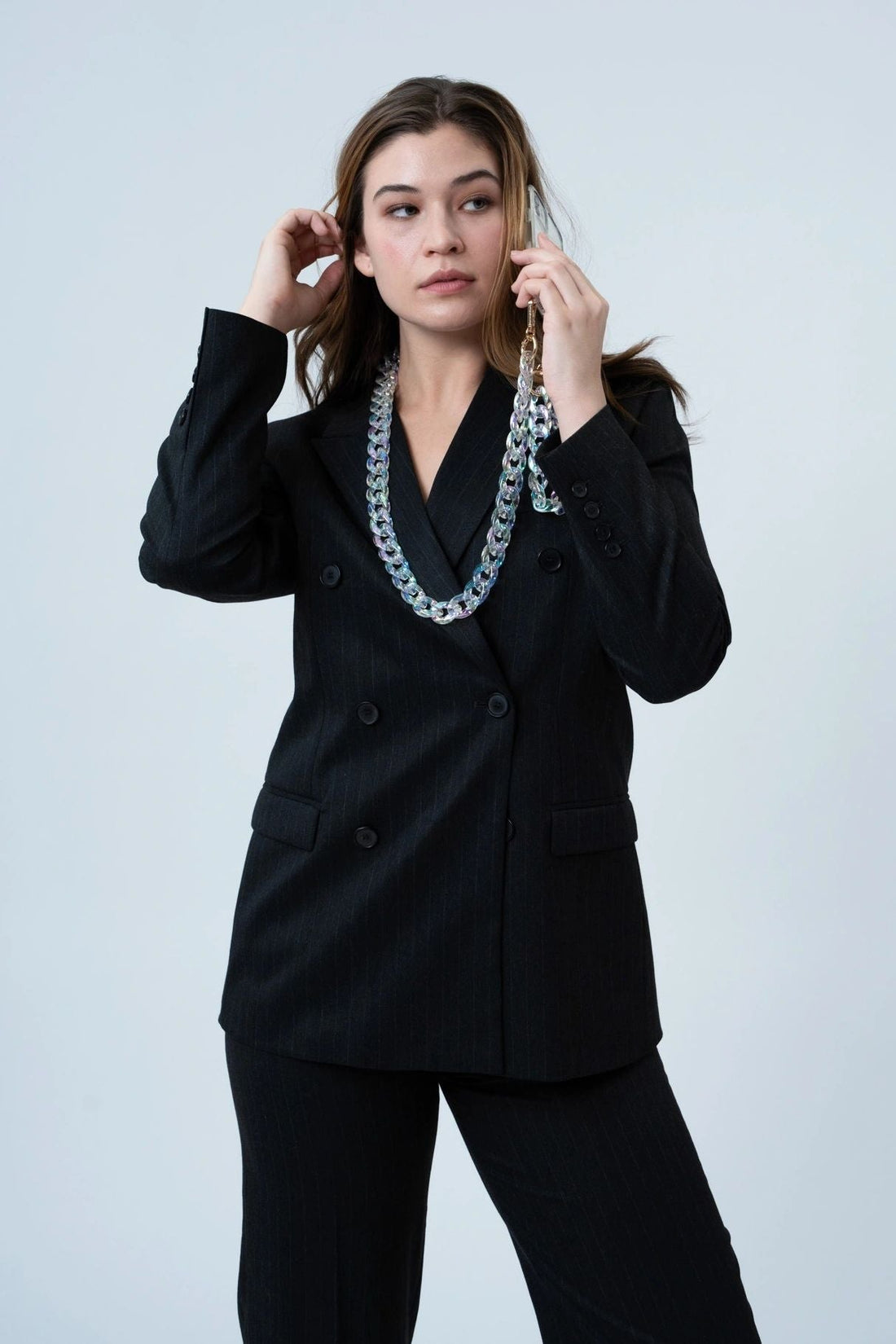 A woman wearing a crystal-effect resin phone chain with gold buckle details, which can be worn as a necklace for added convenience. The chain adds a glamorous touch to her dark jacket. 